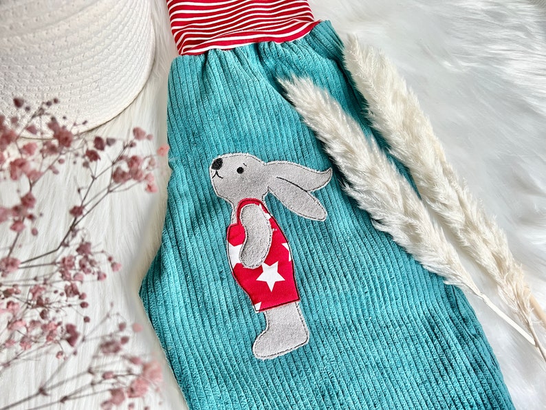 May include: Teal corduroy pants with a red and white striped waistband and a gray bunny applique wearing a red swimsuit with white stars.