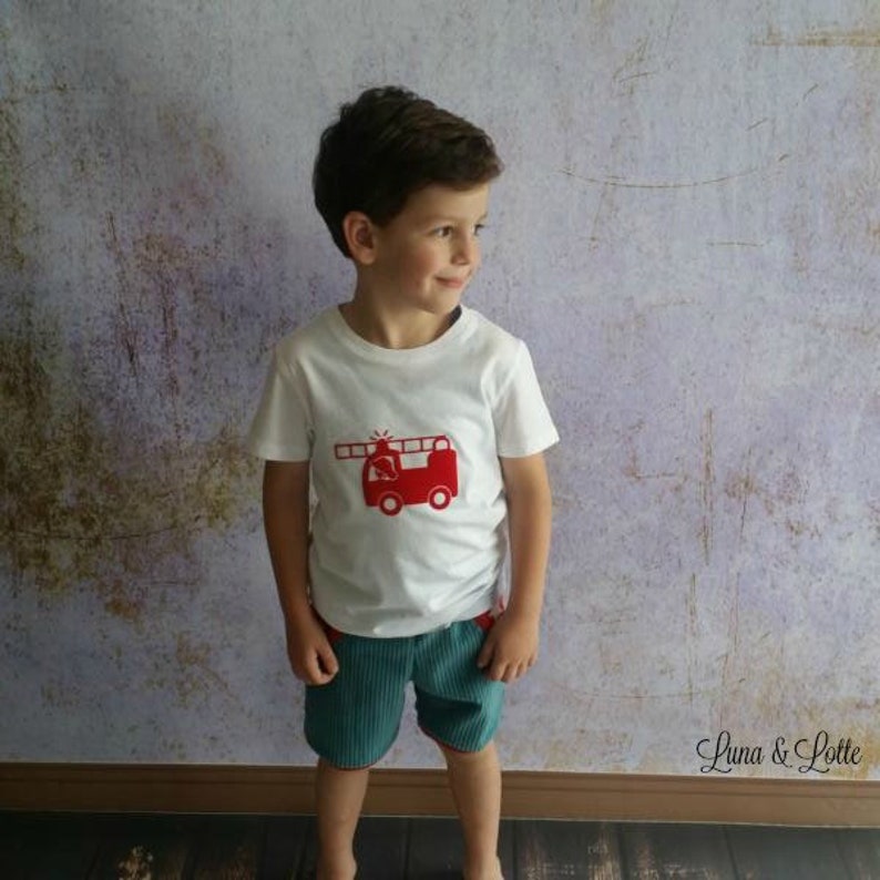 Puede incluir: Una camiseta blanca con un dise&ntilde;o de cami&oacute;n de bomberos rojo, combinada con pantalones cortos de pana turquesa. El dise&ntilde;o del cami&oacute;n de bomberos incluye una escalera y detalles. El conjunto es para ni&ntilde;os.