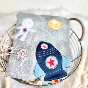 May include: A gray sweatshirt with an astronaut, a planet, and a rocket ship embroidered on it. The sweatshirt is folded and placed in a wire basket.