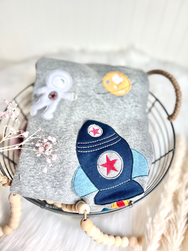 May include: A gray sweatshirt with an astronaut, a planet, and a rocket ship embroidered on it. The sweatshirt is folded and placed in a wire basket.