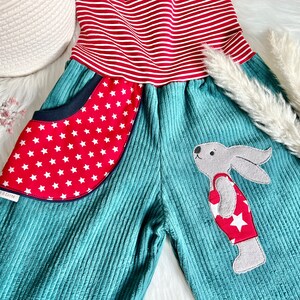 May include: Teal corduroy overalls with red and white striped fabric at the top. The overalls have a red pocket with white stars and a gray bunny applique wearing a red and white star patterned shirt.