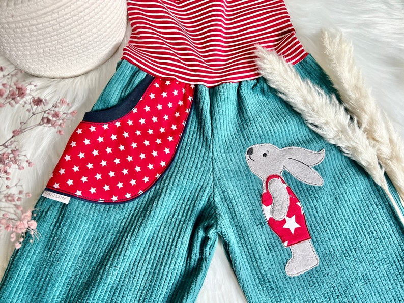 May include: Teal corduroy overalls with red and white striped fabric at the top. The overalls have a red pocket with white stars and a gray bunny applique wearing a red and white star patterned shirt.