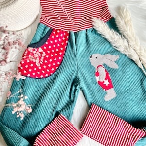 May include: Teal corduroy overalls with red and white striped trim. The overalls have a red pocket with white stars and a patch of a rabbit wearing a red swimsuit with a white star.