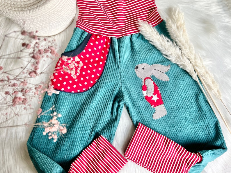 May include: Teal corduroy overalls with red and white striped trim. The overalls have a red pocket with white stars and a patch of a rabbit wearing a red swimsuit with a white star.