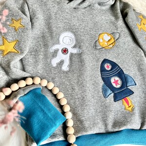 May include: A gray sweatshirt with blue trim features embroidered patches of an astronaut, a rocket, a planet, and stars. The sweatshirt has a hood and long sleeves.