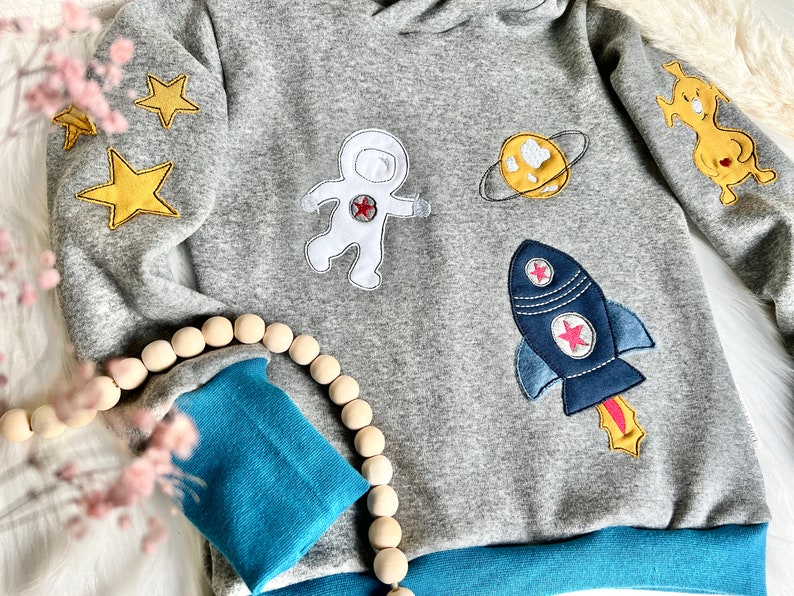 May include: A gray sweatshirt with blue trim features embroidered patches of an astronaut, a rocket, a planet, and stars. The sweatshirt has a hood and long sleeves.
