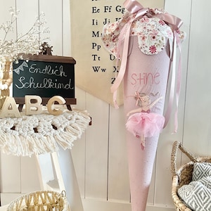 Könnte beinhalten: Eine rosa Schultüte mit floral gemustertem Oberteil und einem Ballerina-Design. Auf der Tüte steht der Name "Stine" und sie ist mit Bändern verziert. Eine Tafel mit den Worten "Endlich Schulkind" ist ebenfalls zu sehen.