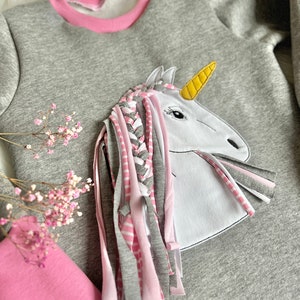 Puede incluir: Una sudadera gris con un ribete rosa alrededor del cuello. Un unicornio blanco con un cuerno amarillo y pelo trenzado rosa y gris est&aacute; aplicado en la parte delantera de la sudadera.