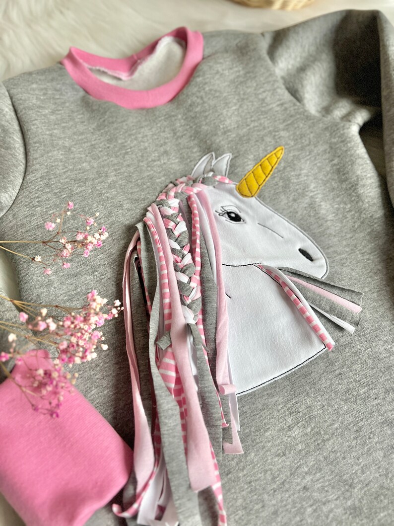 Puede incluir: Una sudadera gris con un ribete rosa alrededor del cuello. Un unicornio blanco con un cuerno amarillo y pelo trenzado rosa y gris est&aacute; aplicado en la parte delantera de la sudadera.