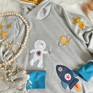 May include: A gray hooded sweatshirt with a blue and yellow trim. The sweatshirt features embroidered astronaut, rocket, planet, and star designs.