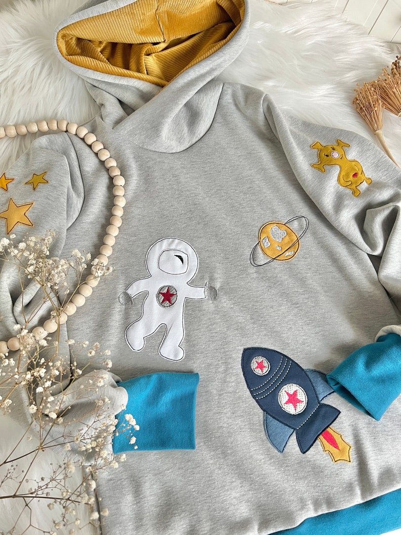 May include: A gray hooded sweatshirt with a blue and yellow trim. The sweatshirt features embroidered astronaut, rocket, planet, and star designs.