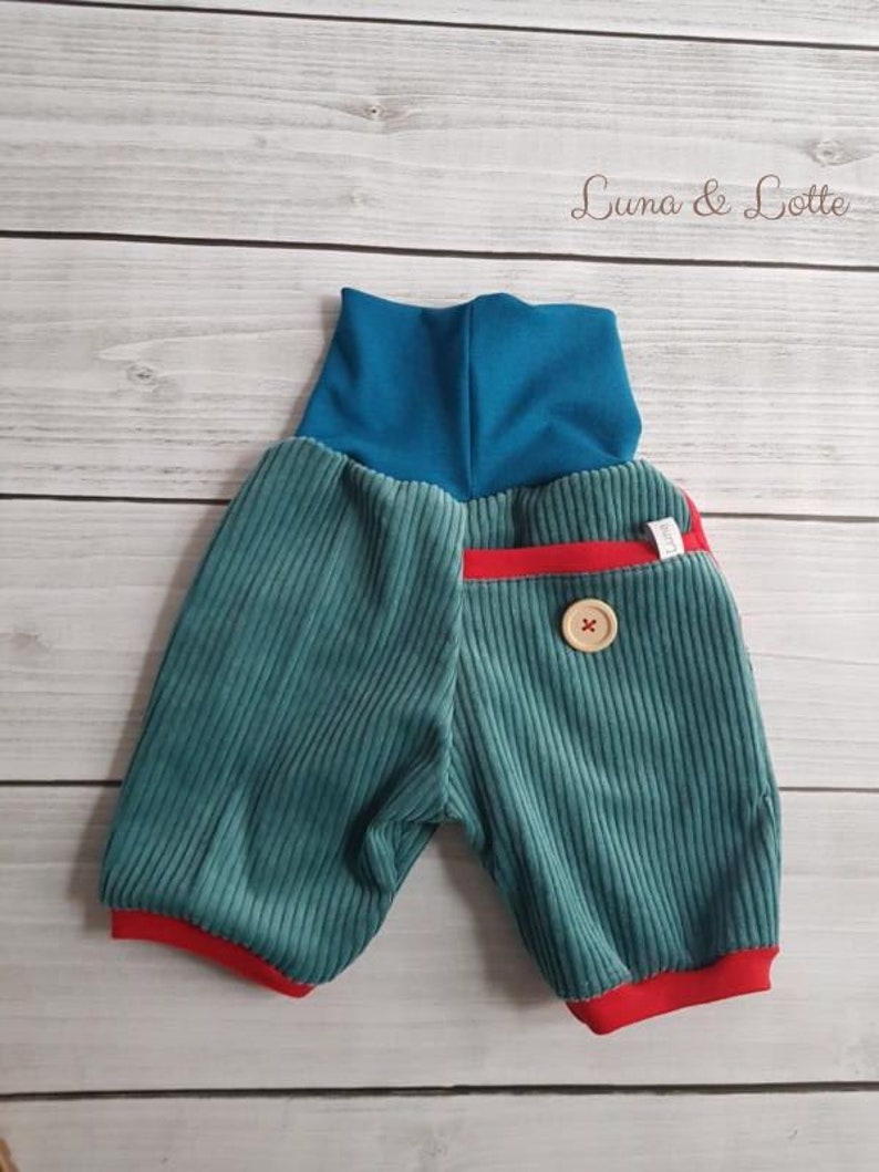 Puede incluir: Shorts de beb&eacute; de pana turquesa con cintura alta turquesa y ribete rojo. Un bot&oacute;n de madera adorna la parte delantera. Los pantalones cortos est&aacute;n etiquetados "Luna & Lotte" en cursiva.