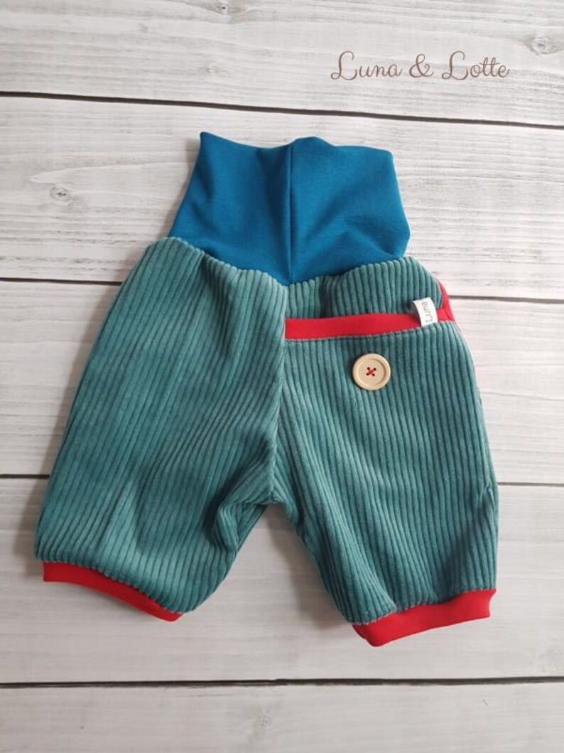Puede incluir: Pantalones cortos de beb&eacute; de pana color turquesa con cintura alta turquesa y ribete rojo. Un bot&oacute;n de madera y un peque&ntilde;o bolsillo rojo detallan los pantalones cortos. Los pantalones cortos est&aacute;n etiquetados como "Luna & Lotte".