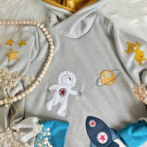 Könnte beinhalten: Ein grauer Kapuzenpullover mit gestickten Astronauten-, Planeten- und Raketenmotiven. Der Pullover hat gelbe Sterne und eine gelbe und graue gestreifte Kapuze.