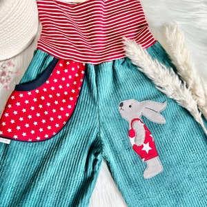 May include: Teal corduroy overalls with a red and white striped waistband and a red pocket with white stars. A gray bunny with a red and white star-patterned shirt is appliqued on the front.
