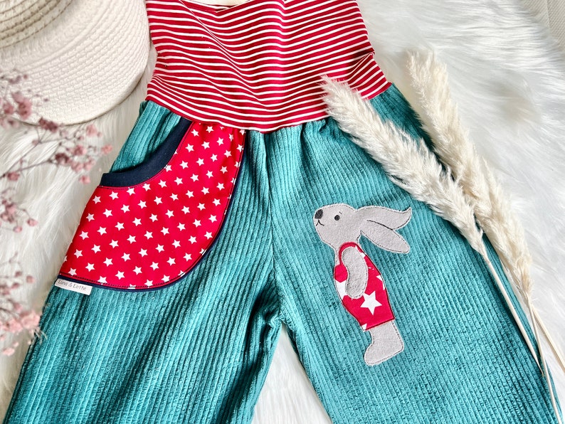 May include: Teal corduroy overalls with a red and white striped waistband and a red pocket with white stars. A gray bunny with a red and white star-patterned shirt is appliqued on the front.