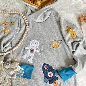 May include: A gray hooded sweatshirt with a blue and yellow trim. The sweatshirt has embroidered space-themed designs, including an astronaut, a rocket, a planet, and stars.