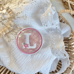 Könnte beinhalten: Ein runder, rosa-weißer Aufnäher mit dem Buchstaben „L“ und einer kleinen Blume. Der Aufnäher befindet sich auf einem weißen, strukturierten Stoff mit weißer Spitzenborte. Im Hintergrund befinden sich ein Weidenkorb und Trockenblumen.
