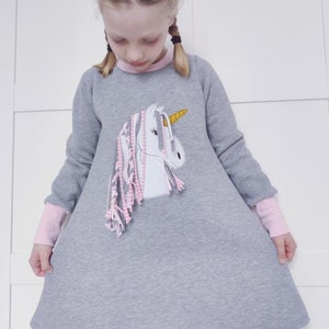Puede incluir: Un vestido de sudadera gris con mangas largas y una aplicaci&oacute;n de unicornio rosa. El unicornio tiene un cuerno dorado y rayas rosas y blancas. El vestido tiene un ribete rosa alrededor del cuello y los pu&ntilde;os.