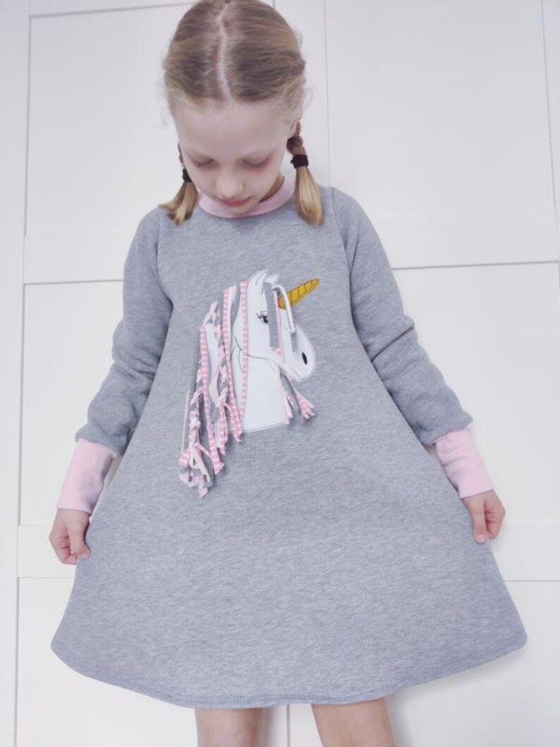 Puede incluir: Un vestido de sudadera gris con mangas largas y una aplicaci&oacute;n de unicornio rosa. El unicornio tiene un cuerno dorado y rayas rosas y blancas. El vestido tiene un ribete rosa alrededor del cuello y los pu&ntilde;os.
