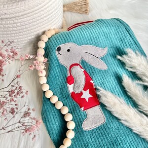 May include: A teal blue corduroy baby blanket with a gray bunny wearing a red swimsuit with white stars. The blanket is surrounded by a wooden bead garland and white pampas grass.