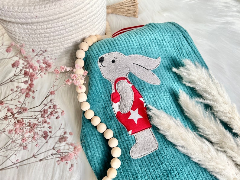 May include: A teal blue corduroy baby blanket with a gray bunny wearing a red swimsuit with white stars. The blanket is surrounded by a wooden bead garland and white pampas grass.