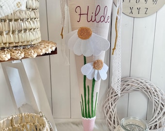 May include: A beige fabric cone-shaped gift bag featuring the name Hilde in dark red script. The bag is decorated with two white felt daisy flowers with yellow centers, green embroidered stems, and a pink fabric base with white lace trim.