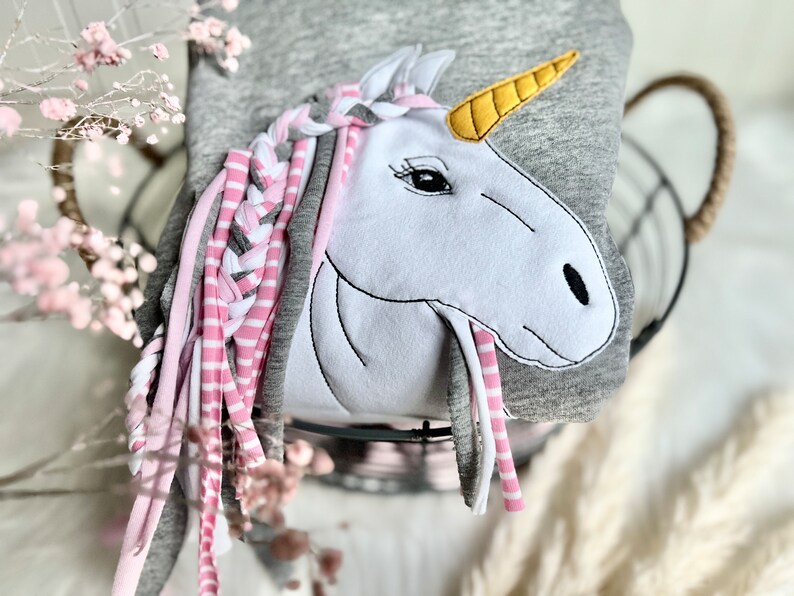 Puede incluir: Una sudadera gris con una aplicaci&oacute;n de unicornio blanco. El unicornio tiene una crin trenzada rosa y gris y un cuerno amarillo.