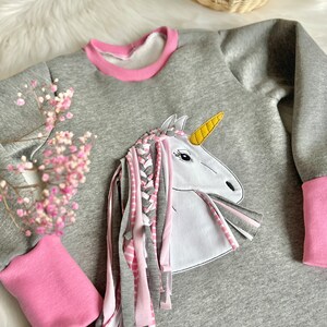 Puede incluir: Sudadera gris con ribete rosa y un aplique de unicornio blanco con un cuerno amarillo y una crin trenzada rosa y blanca.
