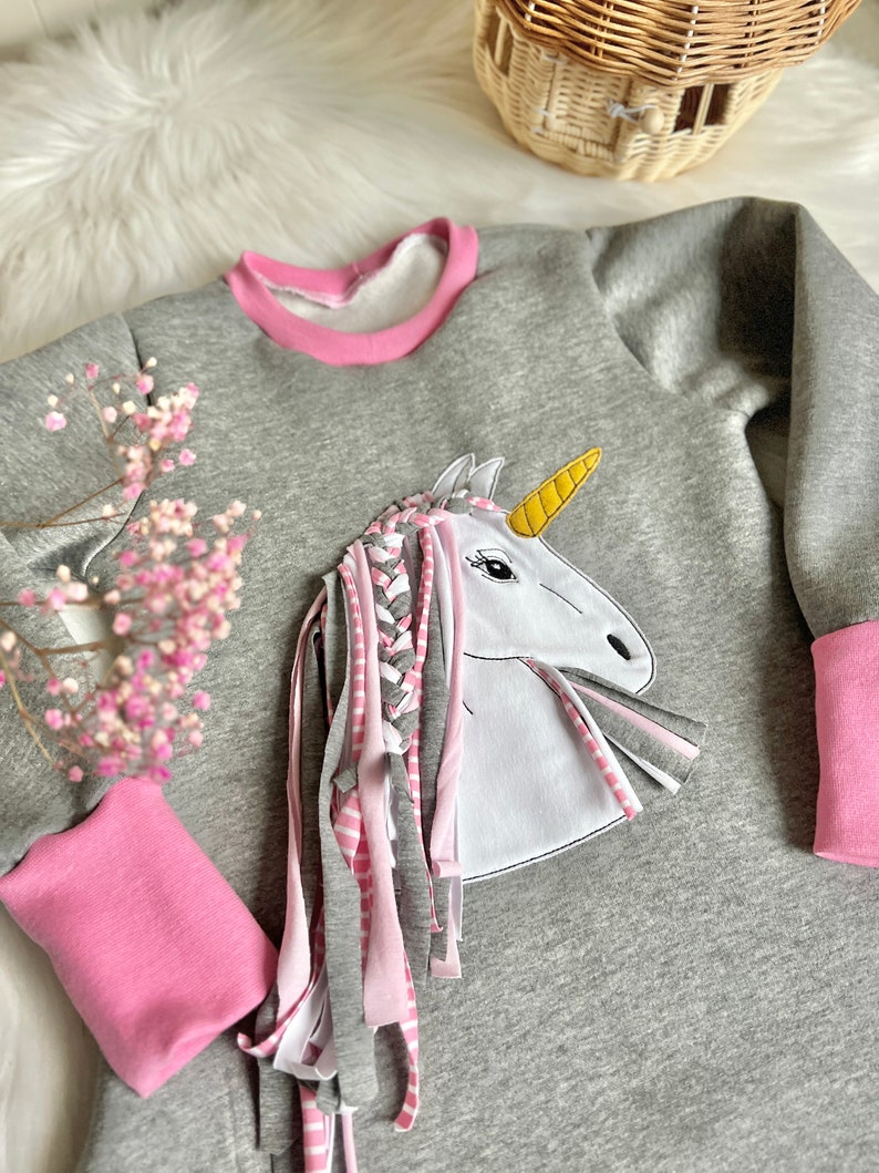 Puede incluir: Sudadera gris con ribete rosa y un aplique de unicornio blanco con un cuerno amarillo y una crin trenzada rosa y blanca.