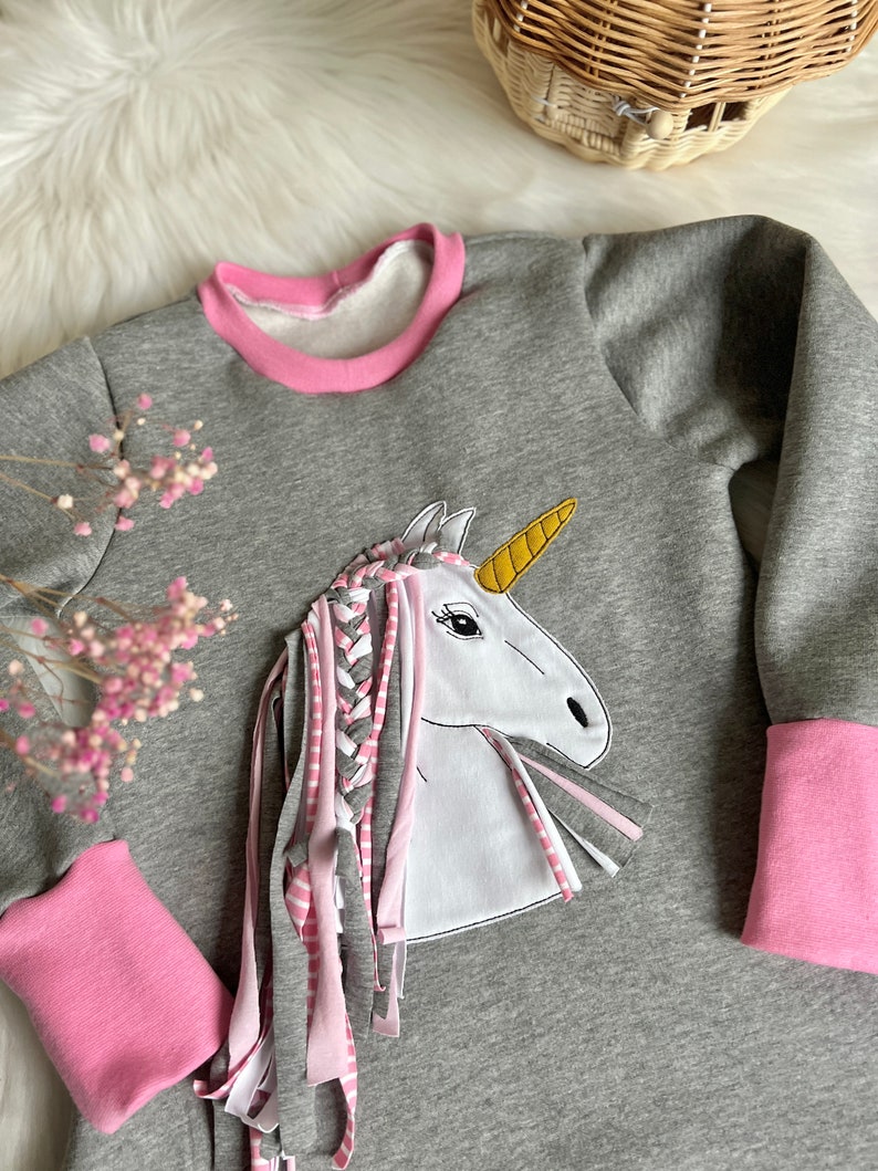 Puede incluir: Sudadera gris con ribete rosa y una aplicaci&oacute;n de unicornio blanco con una crin trenzada y un cuerno dorado. La crin del unicornio est&aacute; hecha de tiras de tela rosa y blanca.