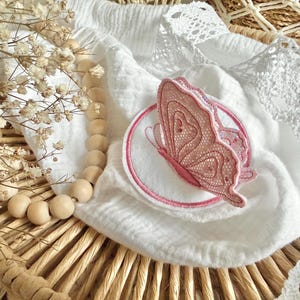Könnte beinhalten: Ein rosa Schmetterlingsapplikation mit filigranen, spitzenartigen Details befindet sich auf einem weißen, kreisförmigen Aufnäher mit rosa Rand. Der Schmetterling ist vor einem weißen, strukturierten Stoff platziert, mit einer Holzperlenkette und Trockenblumen.