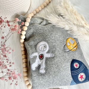May include: Gray sweatshirt with embroidered astronaut, rocket, and planet designs. The astronaut is white with a red star on its chest. The rocket is blue with two red stars. The planet is yellow with a gray ring.