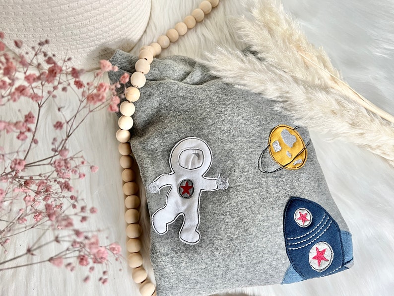 May include: Gray sweatshirt with embroidered astronaut, rocket, and planet designs. The astronaut is white with a red star on its chest. The rocket is blue with two red stars. The planet is yellow with a gray ring.