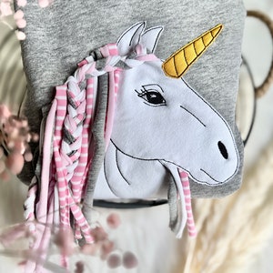 Puede incluir: Una sudadera gris con una aplicaci&oacute;n de unicornio blanco. El unicornio tiene una crin trenzada rosa y gris con mechones largos. El cuerno del unicornio es amarillo.