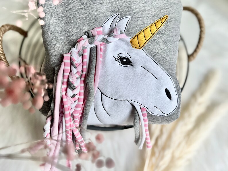 Puede incluir: Una sudadera gris con una aplicaci&oacute;n de unicornio blanco. El unicornio tiene una crin trenzada rosa y gris con mechones largos. El cuerno del unicornio es amarillo.