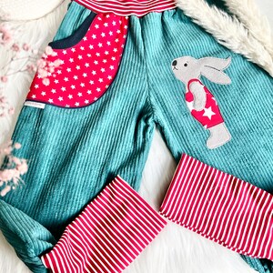 May include: Teal corduroy pants with red and white striped cuffs. The pants have a red pocket with white stars and a felt bunny applique wearing a red and white swimsuit with a white star.