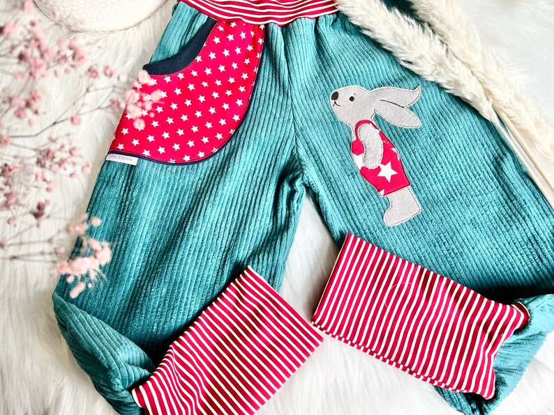 May include: Teal corduroy pants with red and white striped cuffs. The pants have a red pocket with white stars and a felt bunny applique wearing a red and white swimsuit with a white star.