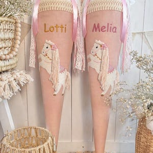 Könnte beinhalten: Zwei rosa Schultüten mit den Namen "Lotti" und "Melia" in Gold gestickt. Jede Tüte zeigt eine weiße Pferdeapplikation mit rosa Blumenkrone und langer Mähne. Die Tüten sind mit Spitze und rosa Bändern verziert.