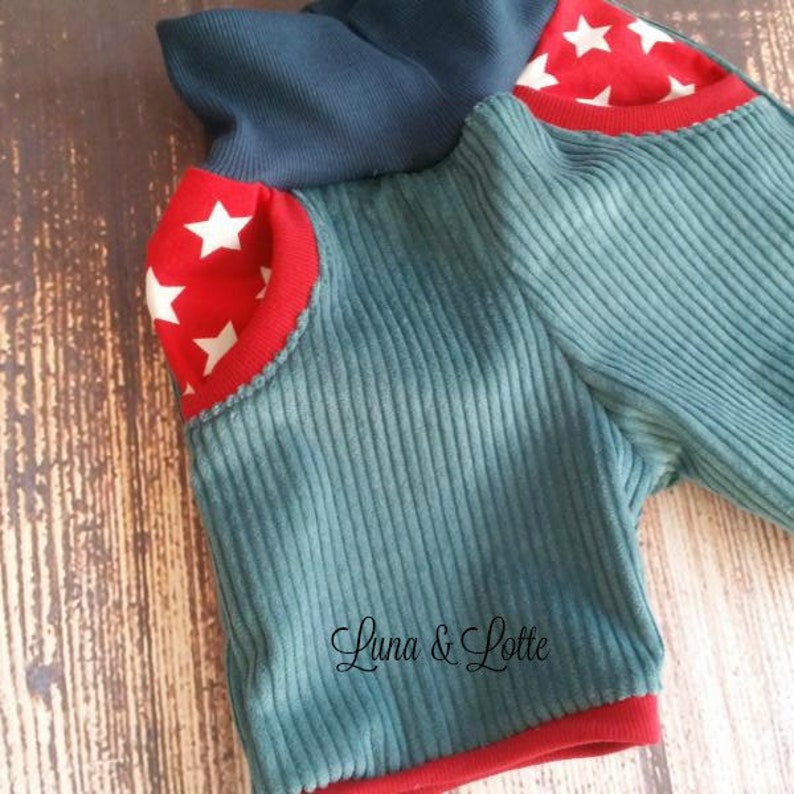Puede incluir: Primer plano de una parte superior de pana color turquesa con ribetes rojos y detalles de estrellas. La parte superior tiene un cuello azul marino y el texto "Luna & Lotte" en cursiva.