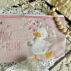 Könnte beinhalten: Eine hellrosa Reißverschlusstasche mit einer gestickten Ente unter einem Regenschirm. Die Ente hat einen gelben Schnabel und Füße. Der Text "TANZ IM REGEN" ist rosa gestickt. Die Tasche hat einen rosa Reißverschluss und Besatz.