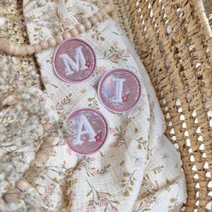 Könnte beinhalten: Drei kreisförmige Aufnäher mit den weißen Buchstaben M, I und A auf einem schimmernden rosa Hintergrund mit kleinen Blumenakzenten. Die Aufnäher sind auf einem cremefarbenen Stoff mit zartem Blumenmuster und Holzperlen angeordnet.
