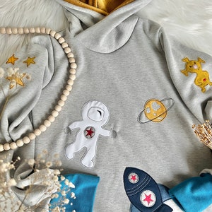 May include: A gray hooded sweatshirt with embroidered space-themed designs, including an astronaut, a planet, and a rocket. The sweatshirt has blue cuffs and a yellow hood.