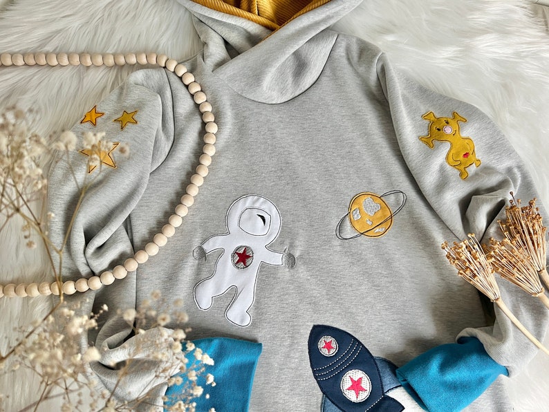 May include: A gray hooded sweatshirt with embroidered space-themed designs, including an astronaut, a planet, and a rocket. The sweatshirt has blue cuffs and a yellow hood.