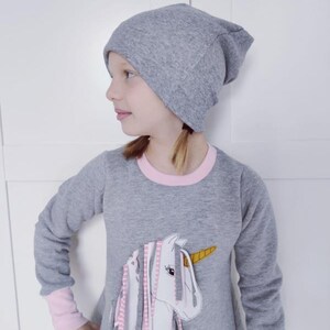 Puede incluir: Un vestido gris de manga larga con un ribete rosa alrededor del cuello y los pu&ntilde;os. El vestido presenta una aplicaci&oacute;n de unicornio blanco con flecos rosa, gris y blanco. La modelo lleva una boina gris.