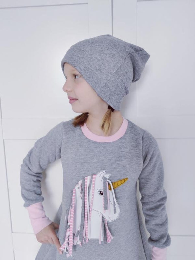 Puede incluir: Un vestido gris de manga larga con un ribete rosa alrededor del cuello y los pu&ntilde;os. El vestido presenta una aplicaci&oacute;n de unicornio blanco con flecos rosa, gris y blanco. La modelo lleva una boina gris.