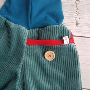 Puede incluir: Primer plano de un pantal&oacute;n de pana verde azulado con un cuello alto verde azulado. El pantal&oacute;n presenta un detalle de bolsillo rojo y un bot&oacute;n grande de color claro con un dise&ntilde;o de cruz. La marca "Luna & Lotte" es visible en la esquina superior derecha.