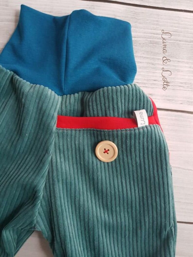 Puede incluir: Primer plano de un pantal&oacute;n de pana verde azulado con un cuello alto verde azulado. El pantal&oacute;n presenta un detalle de bolsillo rojo y un bot&oacute;n grande de color claro con un dise&ntilde;o de cruz. La marca "Luna & Lotte" es visible en la esquina superior derecha.