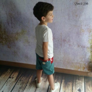 Puede incluir: Un ni&ntilde;o con una camiseta blanca y pantalones cortos de pana verde azulado con forro de bolsillo a cuadros rojos y blancos. Los pantalones cortos tienen un detalle de bot&oacute;n. El ni&ntilde;o lleva zapatillas blancas. El fondo es una pared texturizada y suelo de madera.