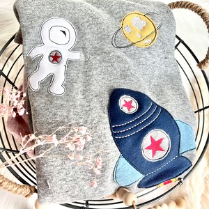 May include: A gray sweatshirt with an astronaut, rocket, and planet applique. The astronaut is white with red and black details. The rocket is blue with red stars and white accents. The planet is yellow with a white heart and a black ring.